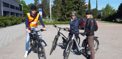 Yksi mies ja kaksi naista seisoo sähköpyörien vieressä kaupunintalon sivustalla.