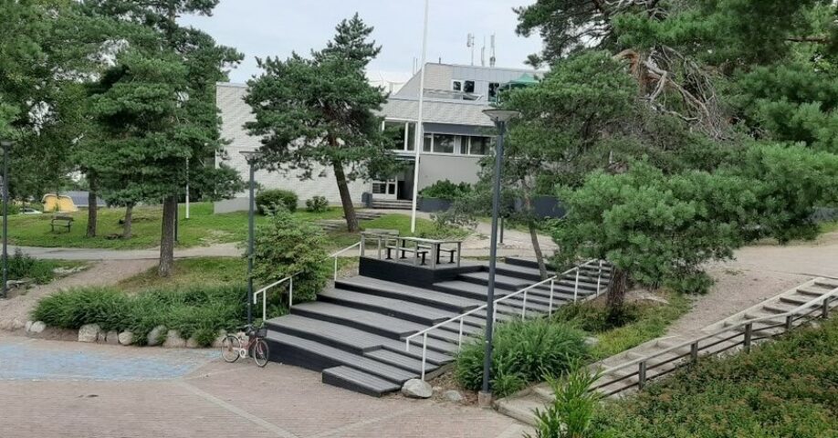 Koulurakennus taustalla, piha-alue ja kiveystä sekä kasvillisuutta edessä
